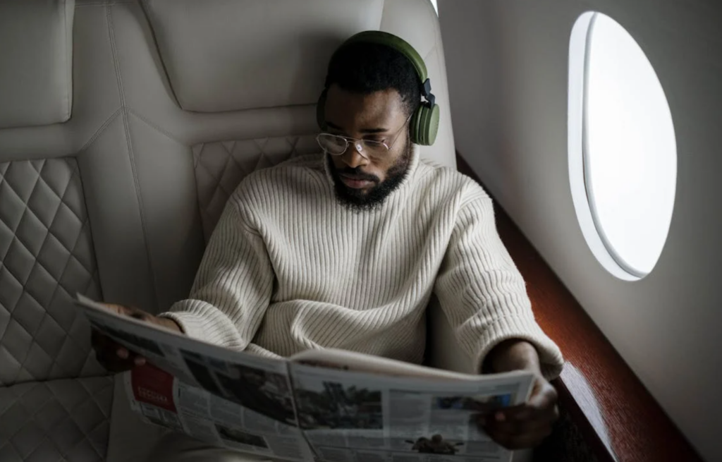 A man relaxing in a business class seat, reading a newspaper with noise-canceling headphones during a flight.
