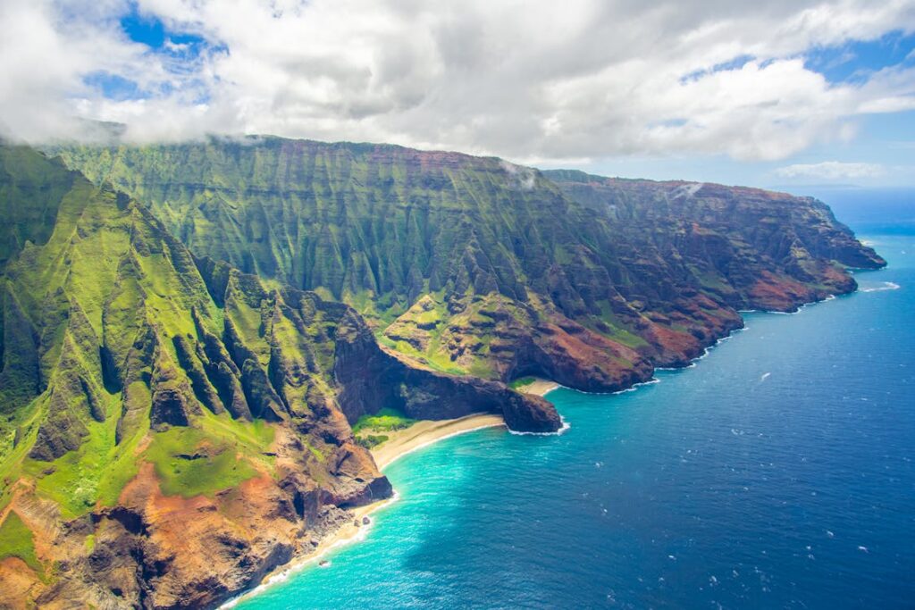 A breathtaking aerial view of the Na Pali Coast in Kauai, Hawaii, showcasing lush green cliffs, turquoise waters, and a secluded sandy beach under a partly cloudy sky.