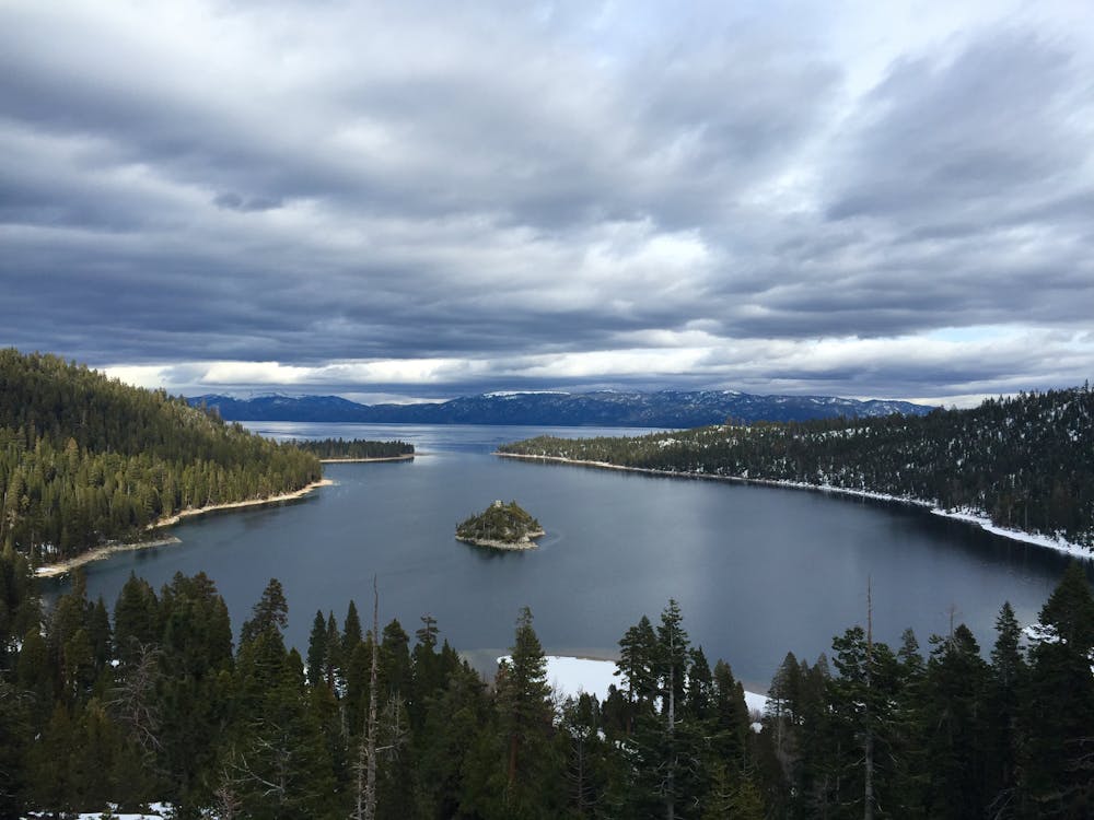A breathtaking view of Lake Tahoe with an island in the center, surrounded by dense evergreen forests and snow-capped mountains under a cloudy sky.
