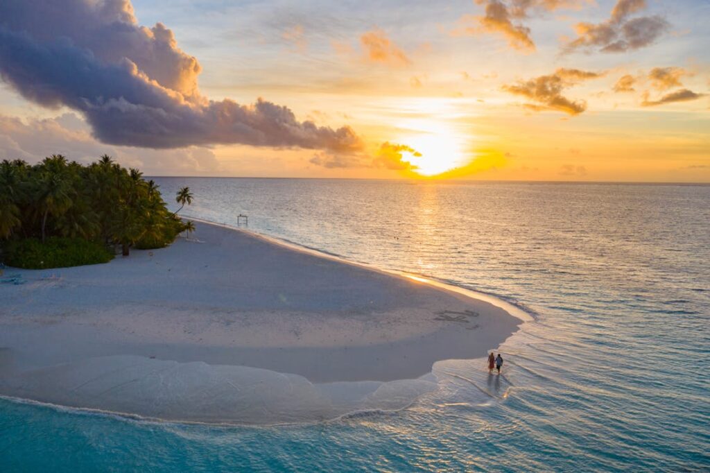 A breathtaking sunset over a secluded sandbank in the Maldives, with a couple walking along the shoreline, surrounded by crystal-clear waters.