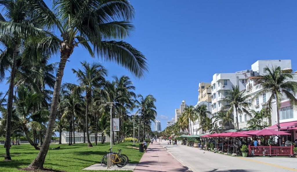 A sunny day at a palm tree-lined pedestrian walkway in South Beach, Miami, with vibrant cafes and white Art Deco buildings on the right.