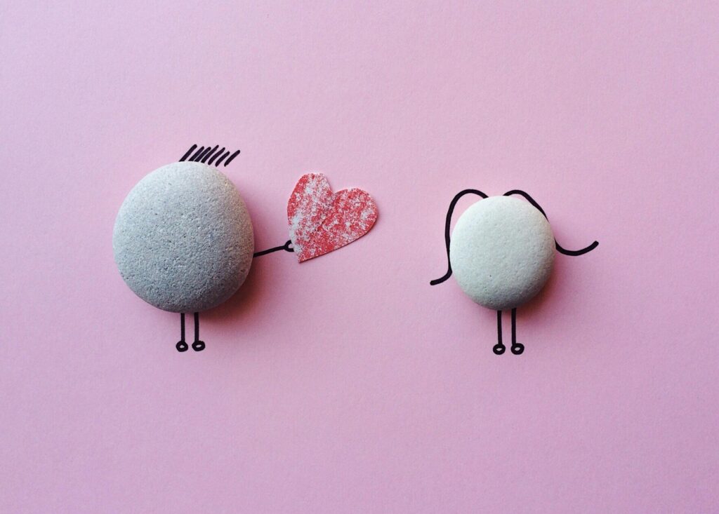Two pebble characters on a pink background, one offering a red heart to the other, symbolizing love and connection.