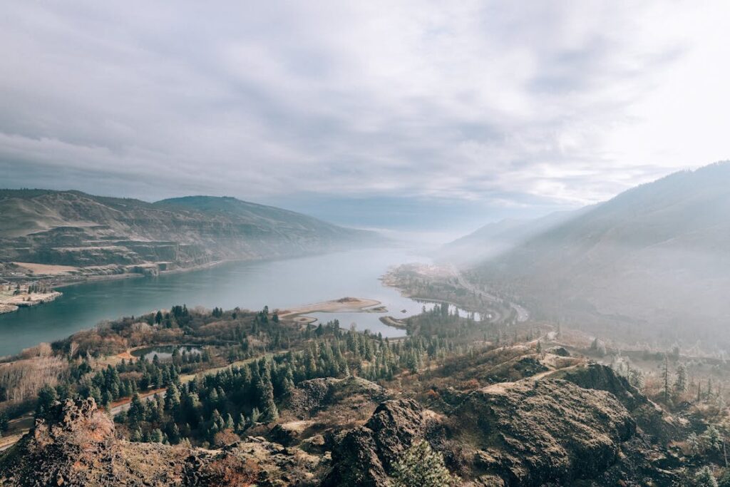 A scenic view of a winding river cutting through lush green hills and mountains under a misty sky in Oregon.