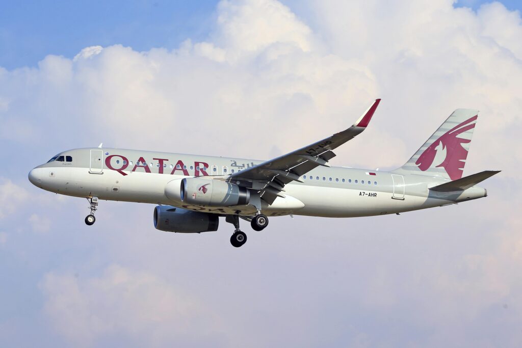 A Qatar Airways Airbus A320 aircraft in mid-flight, approaching for landing against a backdrop of blue skies and white clouds.