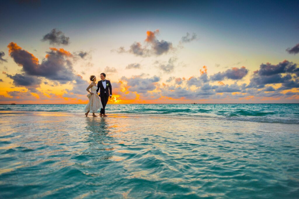 A newlywed couple walks along a shallow beach at sunset, dressed in elegant wedding attire, with the ocean reflecting the golden hues of the sky.