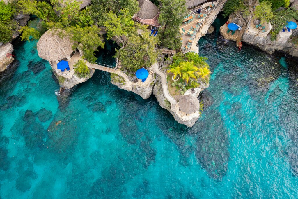 A stunning aerial view of a luxury cliffside resort in Negril, Jamaica, featuring thatched-roof villas, lush greenery, turquoise waters, and private lounge areas with blue umbrellas. A paradise getaway with breathtaking ocean views.