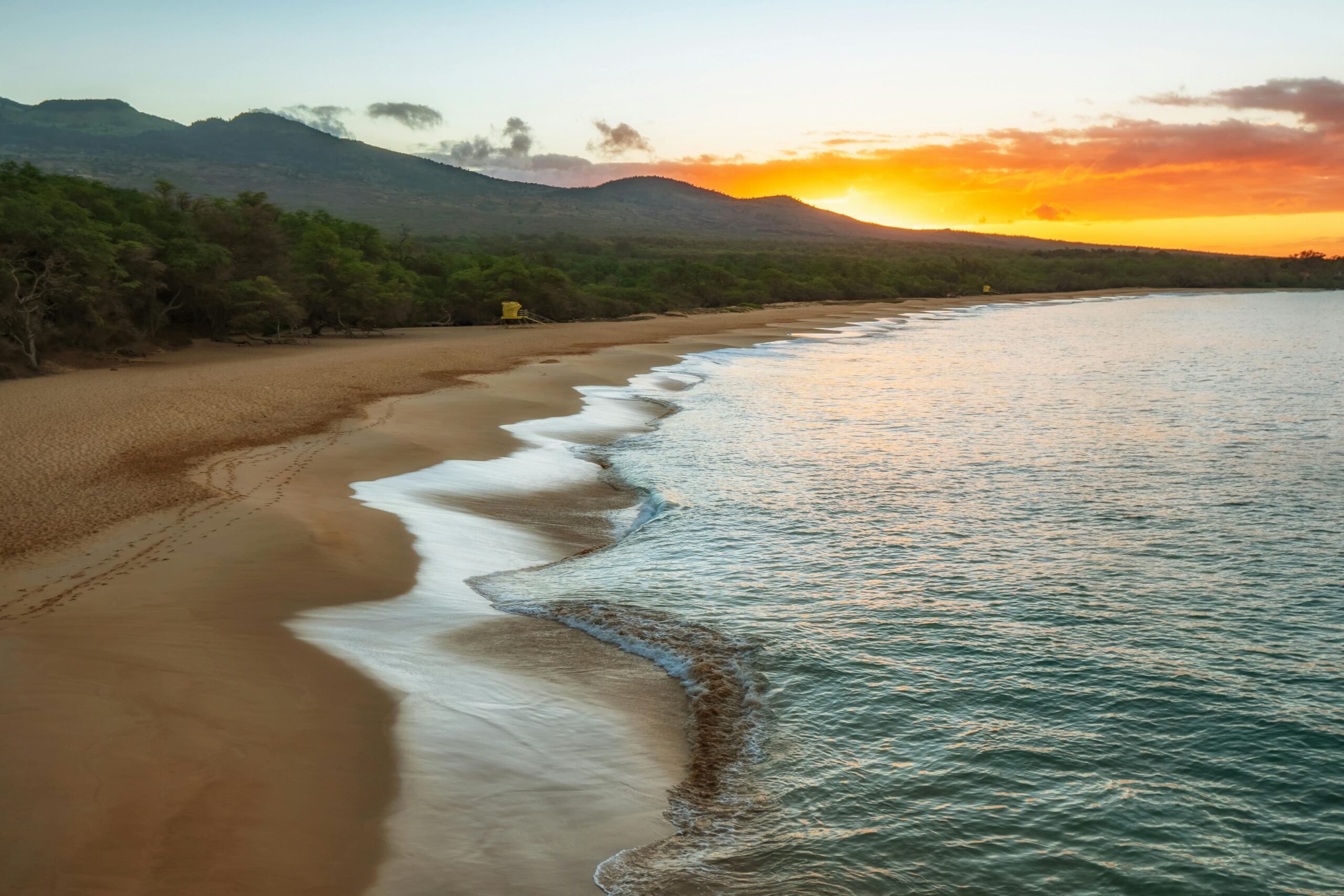 A stunning sunset over a secluded beach in Hawaii, with gentle waves washing onto the golden sand and lush green hills in the background.
