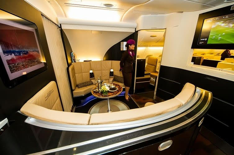 A luxurious onboard lounge in the Etihad A380 Business Class cabin, featuring a circular leather seating area, mounted flat-screen TVs, and a uniformed flight attendant standing nearby. A table in the center displays gourmet refreshments and glassware, highlighting the premium in-flight experience.