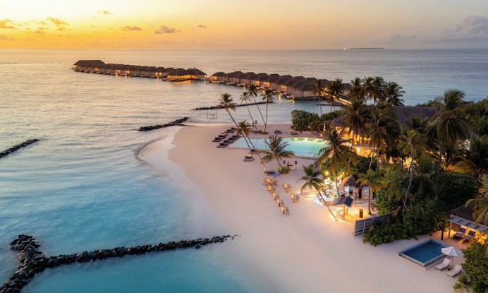 Aerial view of Baglioni Resort Maldives at sunset, showcasing overwater villas, private beach, and glowing pool area.