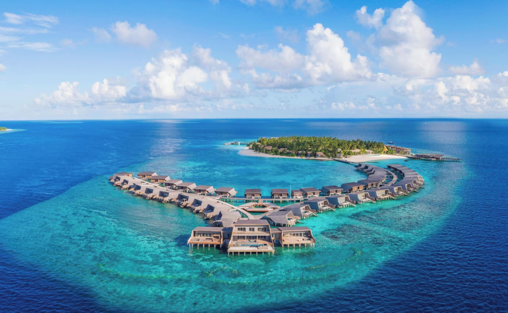 Aerial view of The St. Regis Maldives Vommuli Resort, showcasing overwater villas arranged in a curved formation above turquoise waters with a lush private island in the background.
