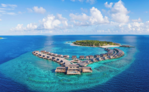 Aerial view of The St. Regis Maldives Vommuli Resort, showcasing overwater villas arranged in a curved formation above turquoise waters with a lush private island in the background.