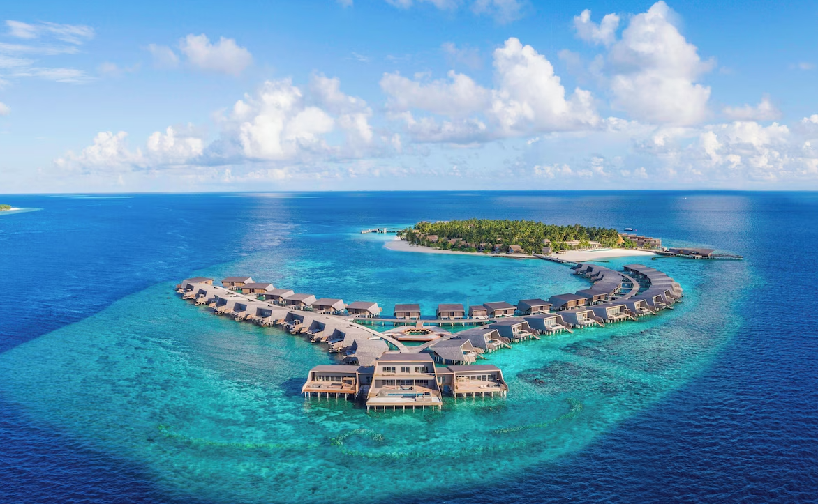 Aerial view of The St. Regis Maldives Vommuli Resort, showcasing overwater villas arranged in a curved formation above turquoise waters with a lush private island in the background.