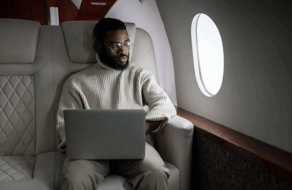 A man wearing headphones and a beige sweater works on a laptop while seated in a luxurious business class airline seat, gazing out the airplane window
