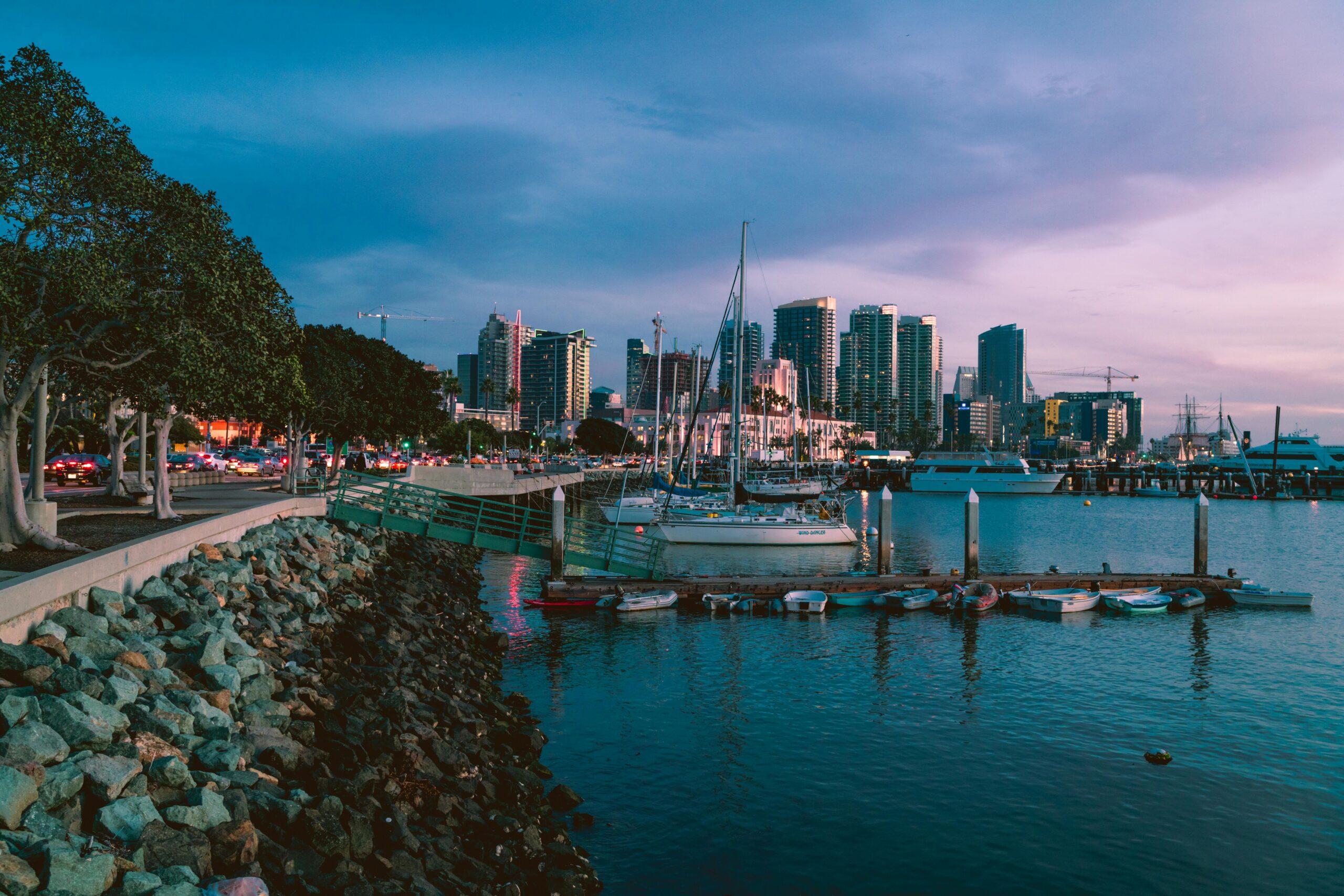 San Diego skyline at sunset with boats docked in the marina and downtown high-rises in the background, viewed from the waterfront near the Gaslamp Quarter.