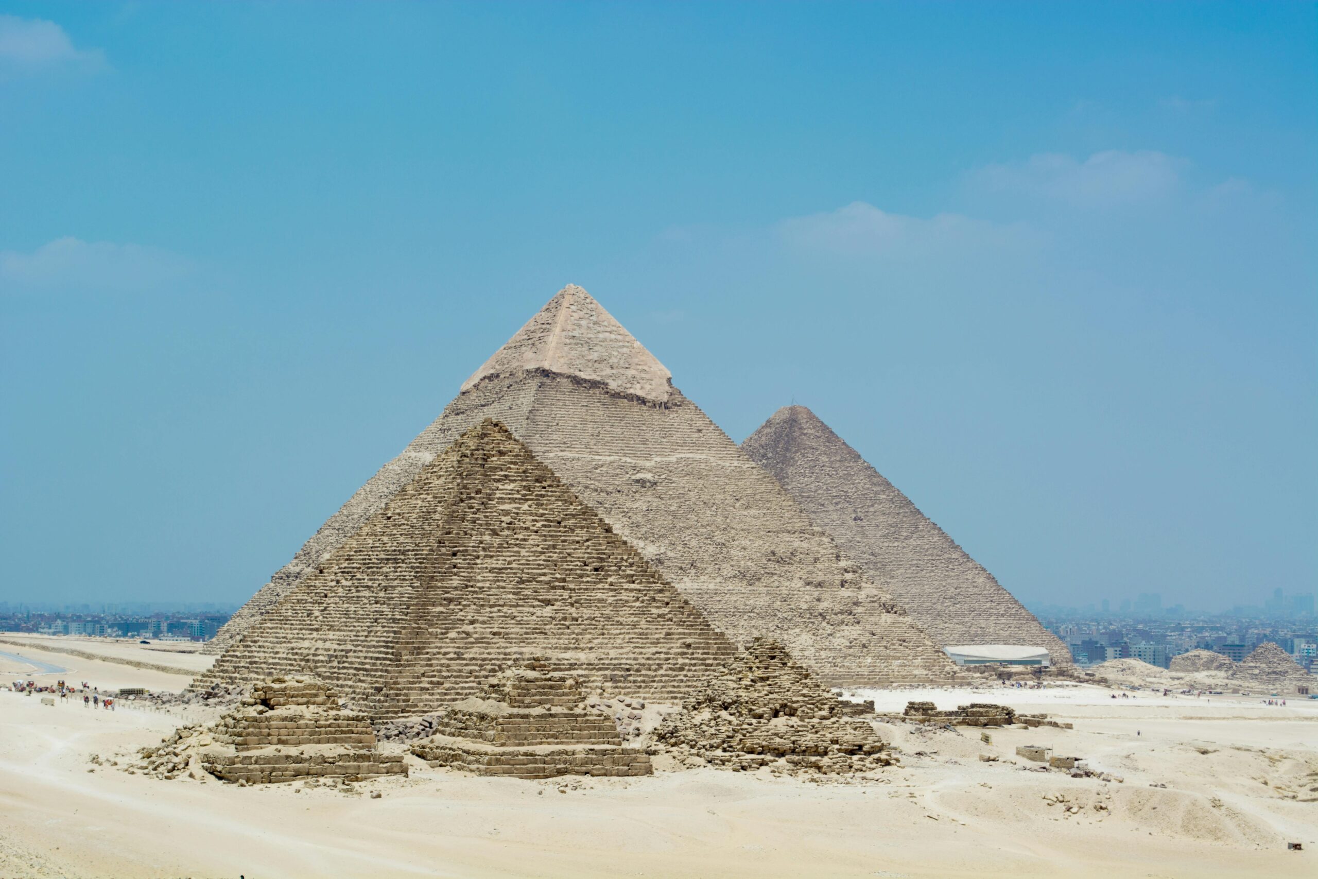 The Great Pyramids of Giza standing tall under a clear blue sky, with the modern Cairo skyline faintly visible in the background.