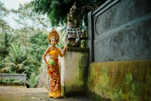 A Balinese woman in traditional ceremonial attire stands beside a stone statue in a lush temple setting.
