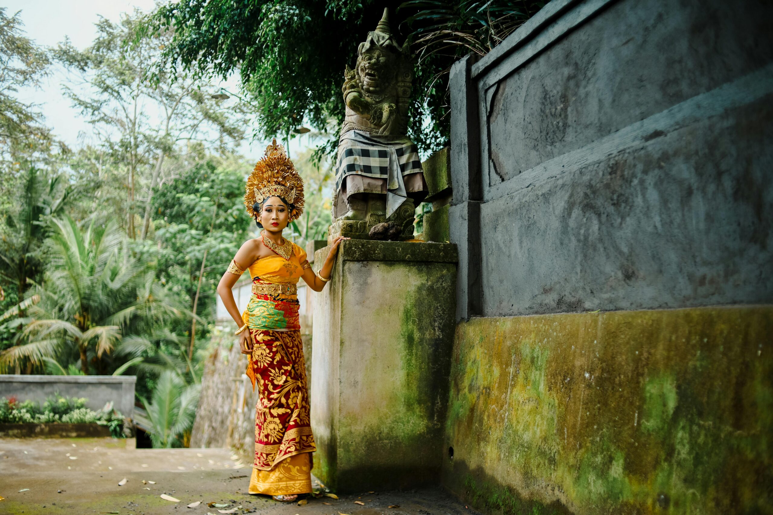 A Balinese woman in traditional ceremonial attire stands beside a stone statue in a lush temple setting.