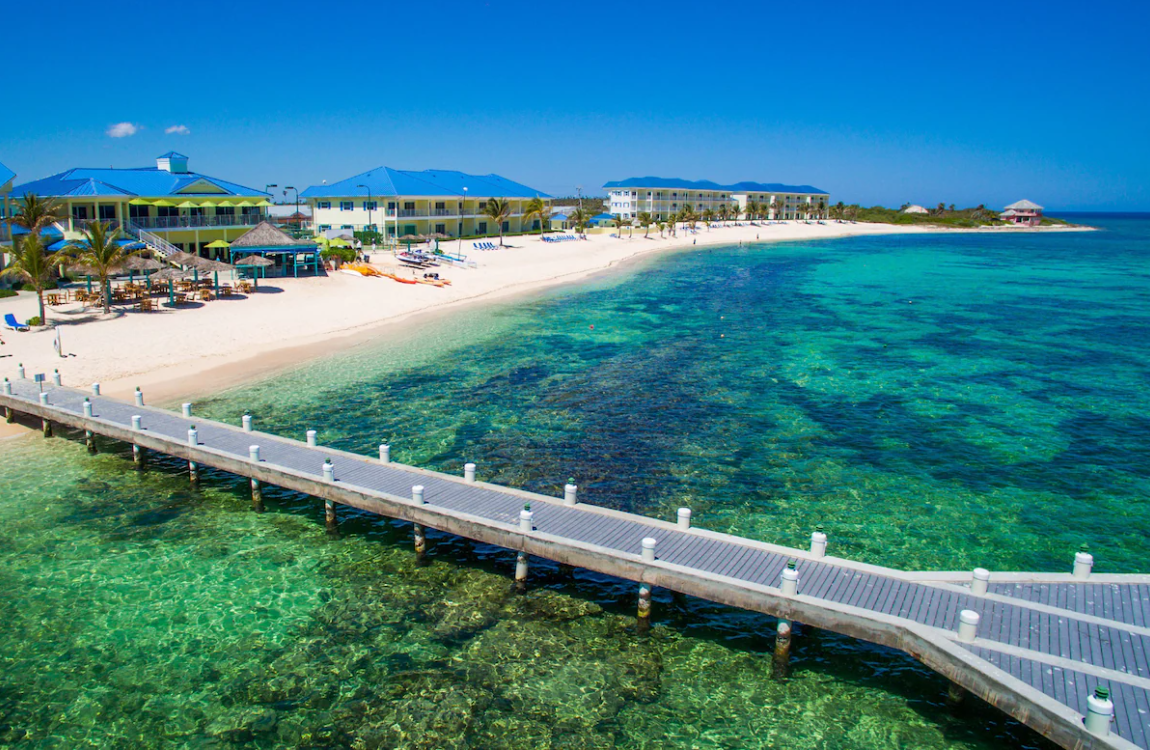 Aerial view of The Reef Beach Resort in East End, Grand Cayman featuring crystal-clear turquoise Caribbean water, white sand beachfront, and oceanfront resort buildings along a private pier.